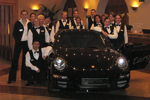 Eine festlich gekleidete Gruppe von Servicemitarbeitern steht strahlend um einen glänzenden schwarzen Porsche herum