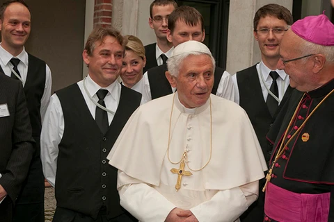 Papst Benedikt XVI. in seiner weißen Soutane, umgeben von mehreren Servicekräfte in Anzügen